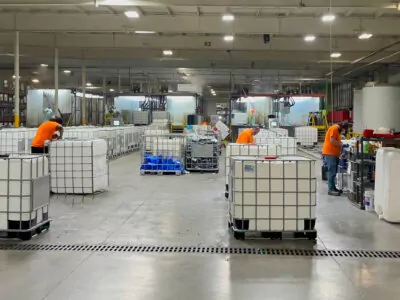 Workers in orange shirts are in a large industrial facility, handling large white containers on pallets. The spacious room is well-lit, with various equipment and shelving in the background. Signs and safety gear are visible throughout.