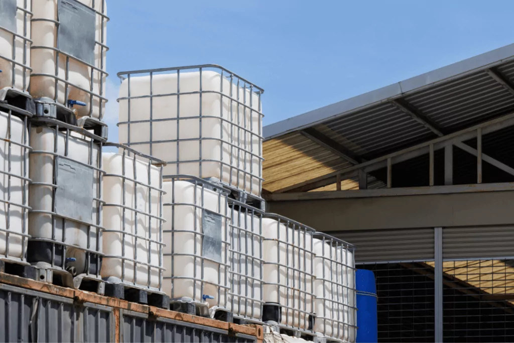 Stacks of large, white industrial containers with metal frames are placed outside near a building with a slanted, corrugated metal roof. The sky is clear and blue.