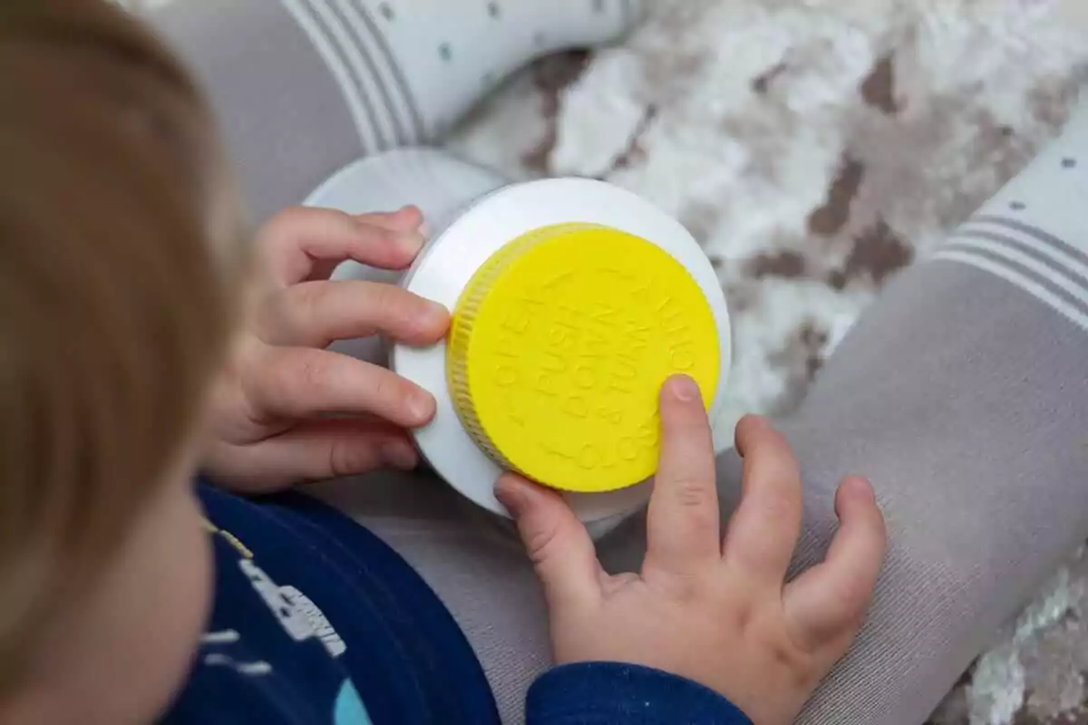 A child with light brown hair is clutching a container featuring child-resistant packaging, complete with a yellow safety cap and opening instructions. Dressed in a blue shirt and gray pants, they sit on a beige-patterned carpet.