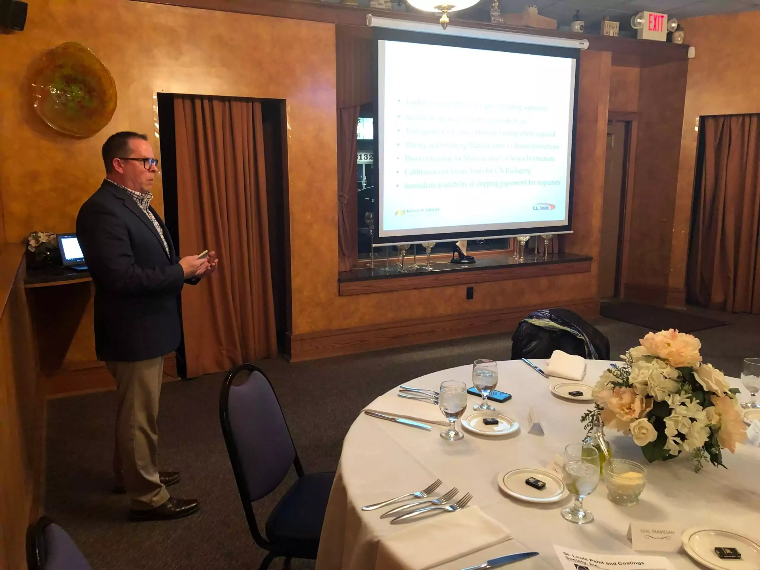 A person from the St. Louis Paint & Coating Society stands next to a presentation screen in a dimly lit room, giving a talk to an unseen audience. A table in the foreground is set for a meal, with a flower centerpiece, plates, cutlery, and glasses.