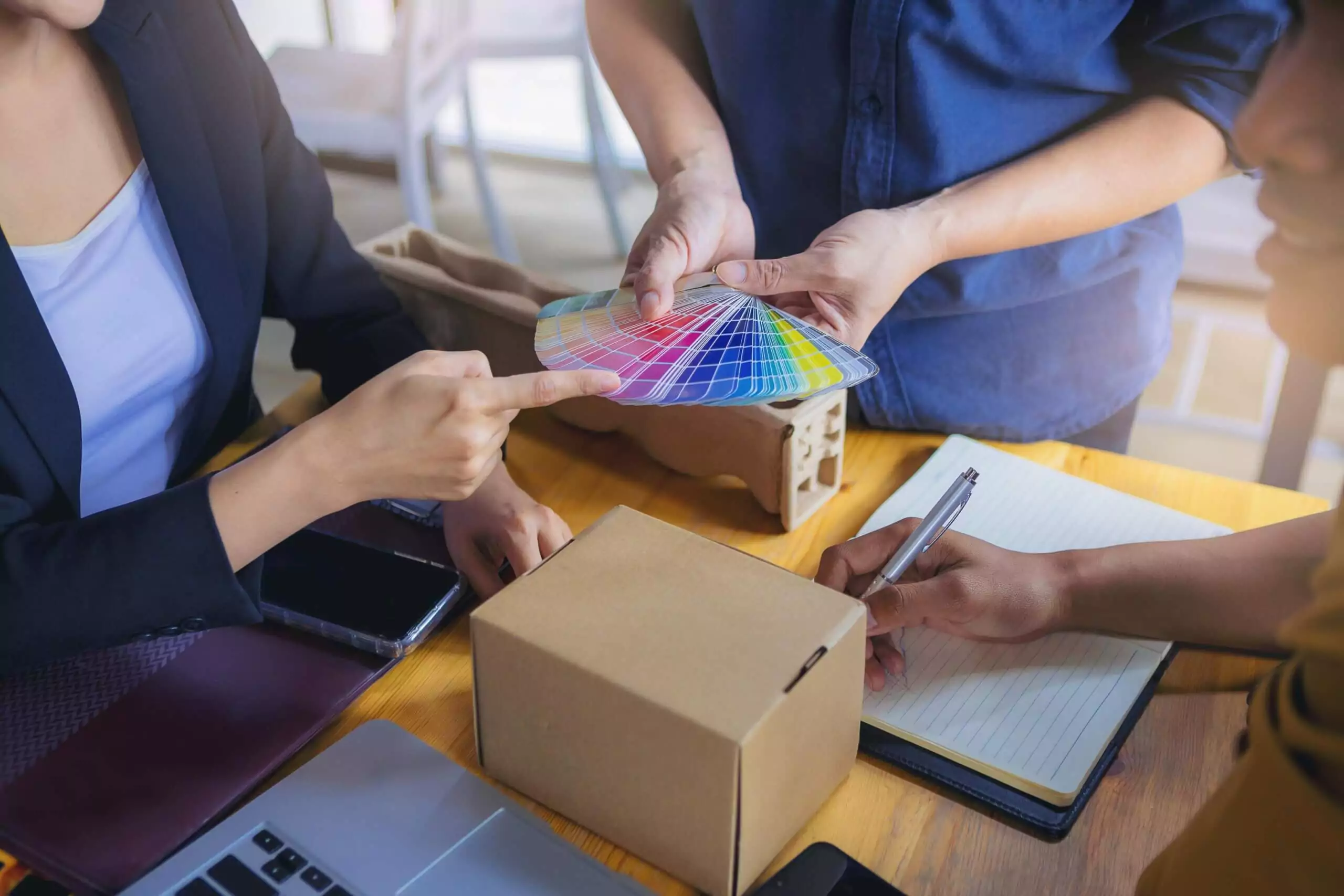 A group of people sits at a table with laptops and notebooks, focused on custom packaging design. One person holds a color palette, another points at it, and a third diligently takes notes next to a small cardboard box.