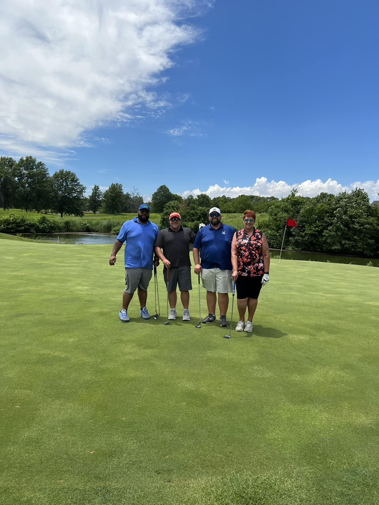 Four participants line up on the lush green of the golf course, clubs in hand, as part of the SLPCS Spring Golf Tournament 2024. Behind them, serene trees and a tranquil pond complement the blue sky dotted with clouds.