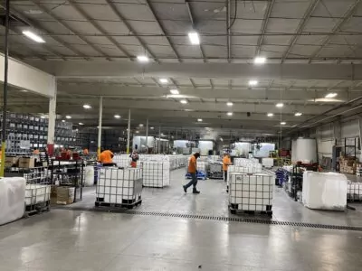 Warehouse interior with several workers in orange shirts moving among large, cube-shaped containers on pallets. Shelves line the walls, and fluorescent lights illuminate the high-ceilinged space.
