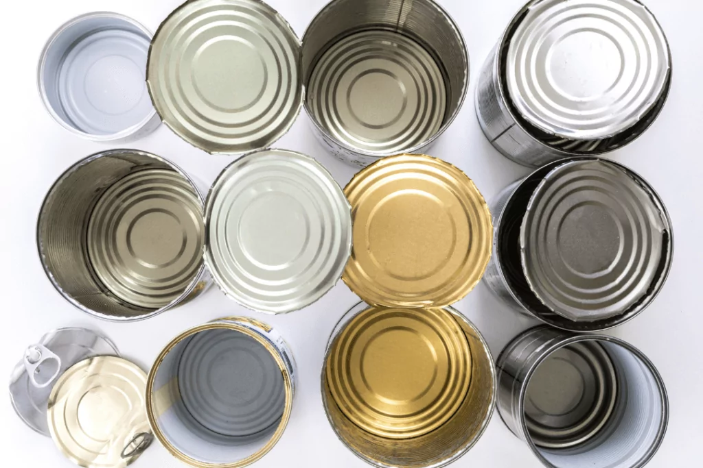 A top view of twelve open, empty metal cans arranged in a grid pattern. The insides are visible, showing different colors such as gold, silver, and gray. The background is a plain light surface.