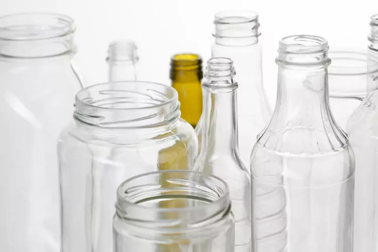 A variety of empty glass jars and bottles in different shapes and sizes are clustered together against a white background. They are mostly clear, with one brown bottle among them.