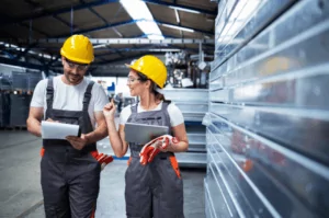 Production Two factory workers in overalls and yellow hard hats are standing in a warehouse. The man is holding a clipboard, and the woman is holding a tablet, discussing services as they converse with metal shelves in the background.