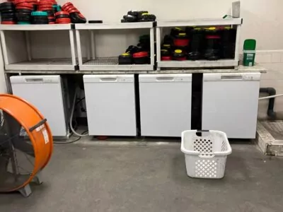 A storage room with three white mini-fridges on the floor. Above them, shelves hold stacks of red and black circular weights. A large orange industrial fan is on the left, and a white laundry basket is on the floor.