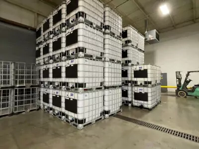 Stacked industrial containers in a warehouse, organized neatly in rows. A forklift is visible in the background, and the space has a large, open layout with a high ceiling and concrete floor.