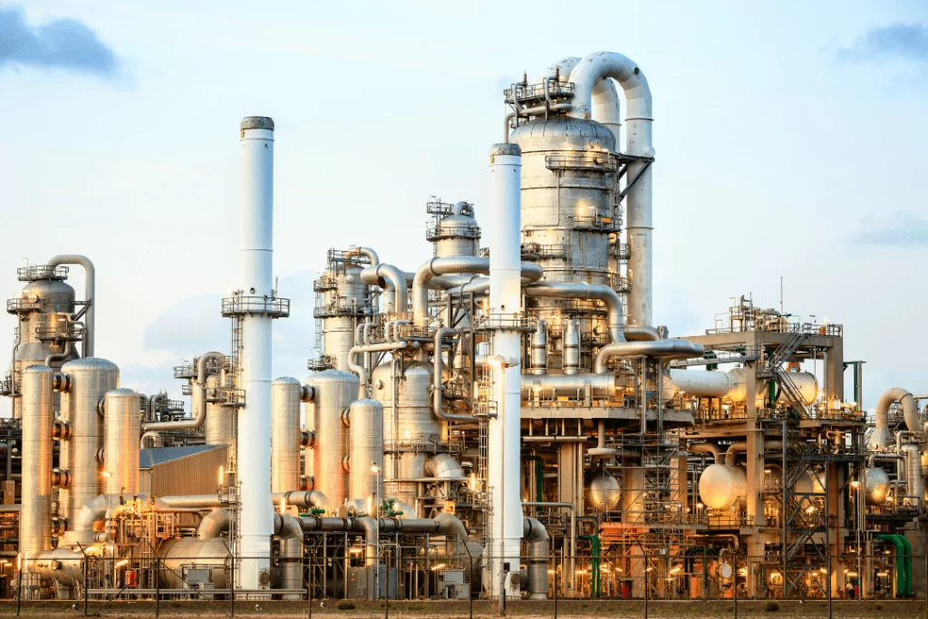Industrial chemical plant with numerous interconnected pipes, towers, and storage tanks. The metallic structures glisten under daylight, showcasing a complex network of machinery against a clear sky.