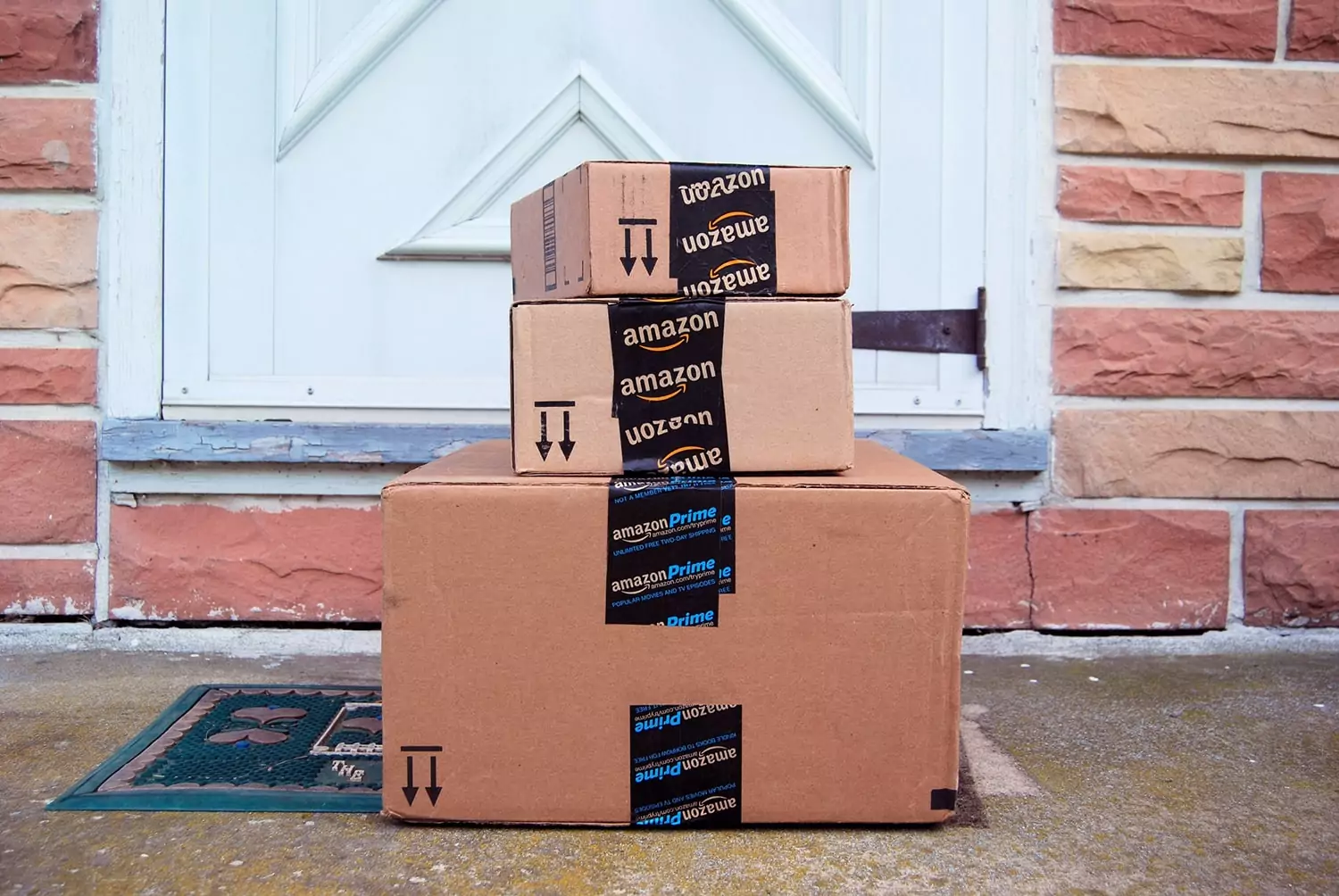 Three stacked Amazon Prime packages are sitting on a doormat in front of a white door with a geometric design. The background features a stone wall.