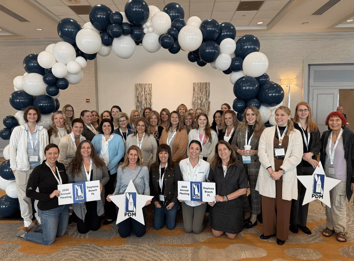 A group of smiling people pose under a blue and white balloon arch indoors. Some are holding signs that read "2023 Employee Reunion" and "PDM." The setting is a well-lit room with carpeted flooring.