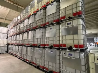 Rows of large, stacked industrial containers in a warehouse with high ceilings. The containers are gray with red tops and supported by metal frames. A concrete floor and shelves in the background are visible.