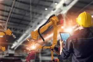Manufacturing A worker in a yellow hard hat efficiently operates a robotic arm using a tablet, showcasing advanced services in an industrial setting. The robotic arm emits sparks as it performs welding or metalwork, surrounded by the expansive factory interior with towering metal structures.
