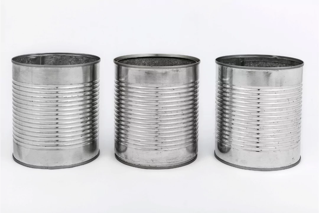 Three empty, silver tin cans with ridged surfaces are aligned side by side against a plain white background.