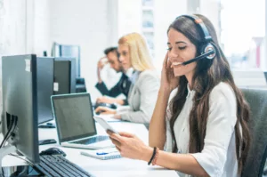 Customer Support A woman with long brown hair, wearing a headset, smiles at her phone while providing services at a desk with computers. In the background, two other people are also wearing headsets and working diligently in a bright office setting.