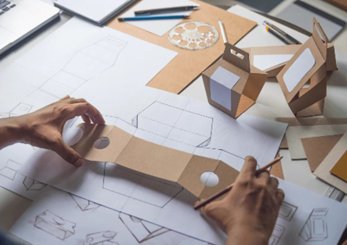 Hands holding and designing a cardboard mock-up on a worktable covered with packaging design sketches, a laptop, and drawing tools.