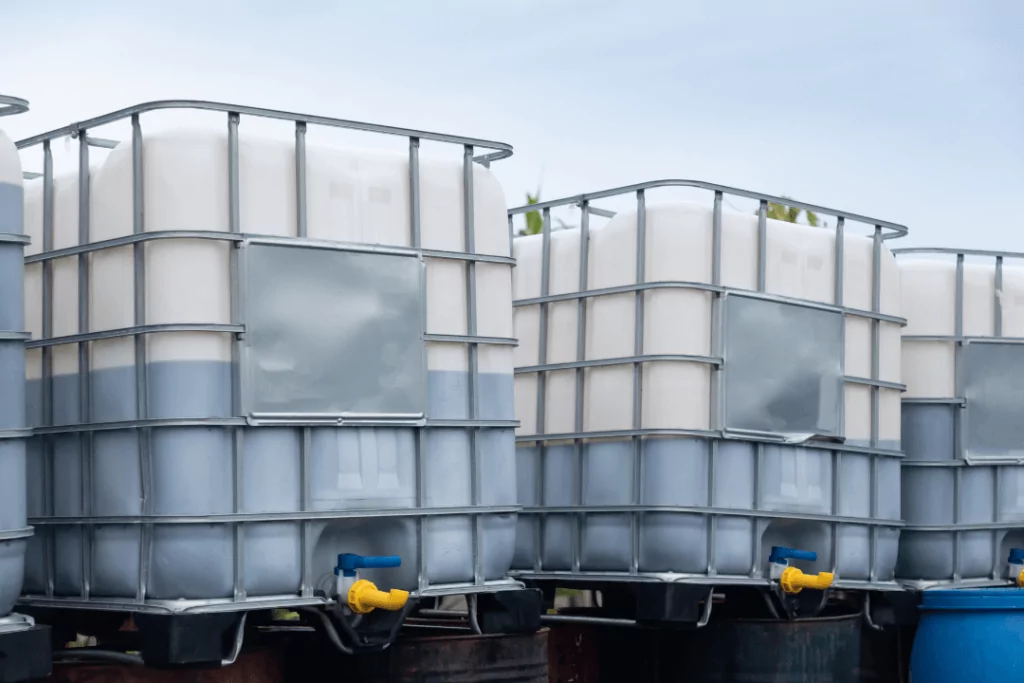 IBC 13 Large industrial containers filled with liquid are stacked outdoors. The containers are white with gray metal cages for support and have blue and yellow spouts at the bottom for dispensing. The sky is overcast in the background.