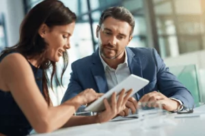 Enhancement A woman and a man are sitting at a desk in a bright office setting, discussing various services. The woman is pointing at a tablet while the man attentively looks at the screen. Both are dressed in business attire, embodying professionalism.