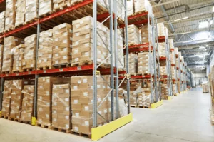 Warehouse Warehouse interior with high shelves filled with stacked, wrapped boxes on pallets. Aisle runs down the middle, with overhead lighting illuminating the scene. The warehouse services ensure a clean and spacious floor, allowing for easy access and movement.