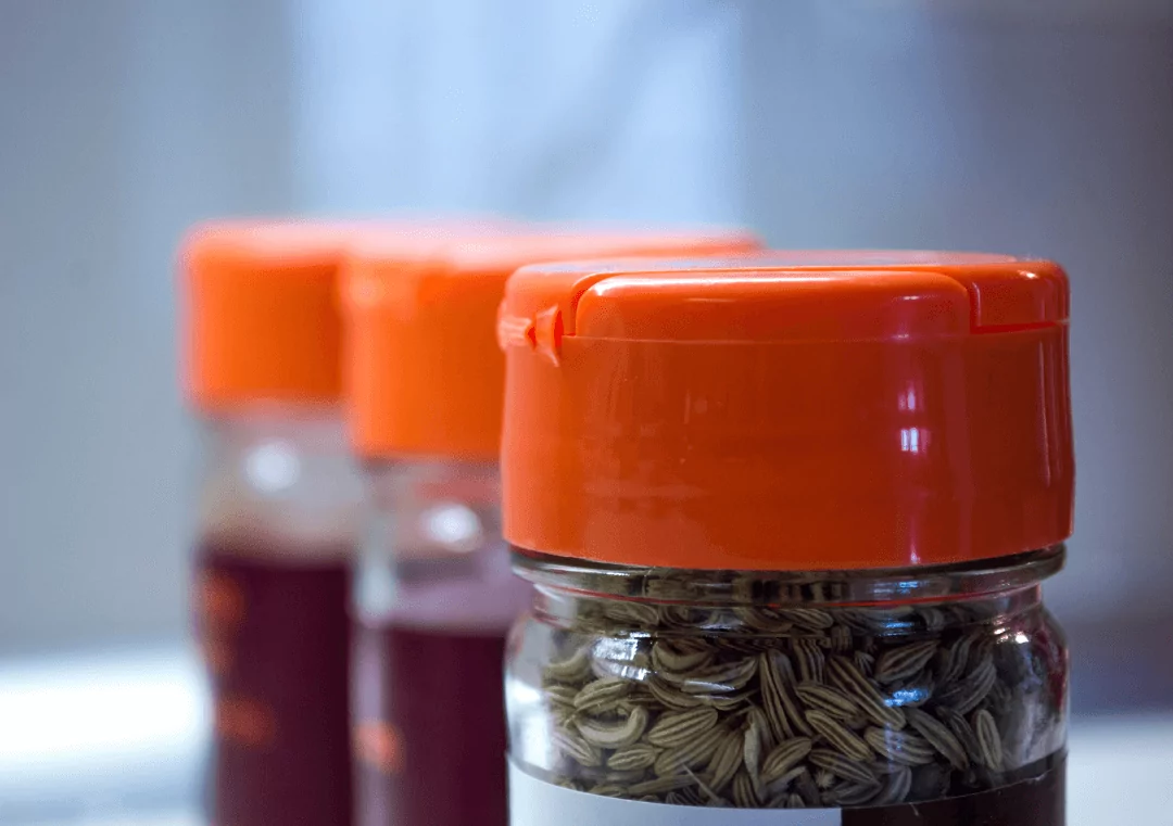Close-up of three spice jars with red-orange lids. The jars are arranged in a row, with the focus on the nearest jar containing fennel seeds. The background is softly blurred, highlighting the vibrant lid color.