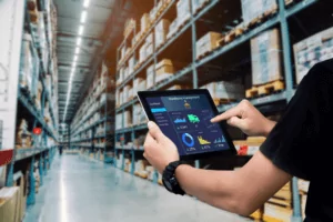 Inventory Management A person using a tablet in a large warehouse filled with shelves stocked with boxes. The tablet displays graphs and data analytics related to inventory management. The scene conveys a sense of modern logistics and technology.
