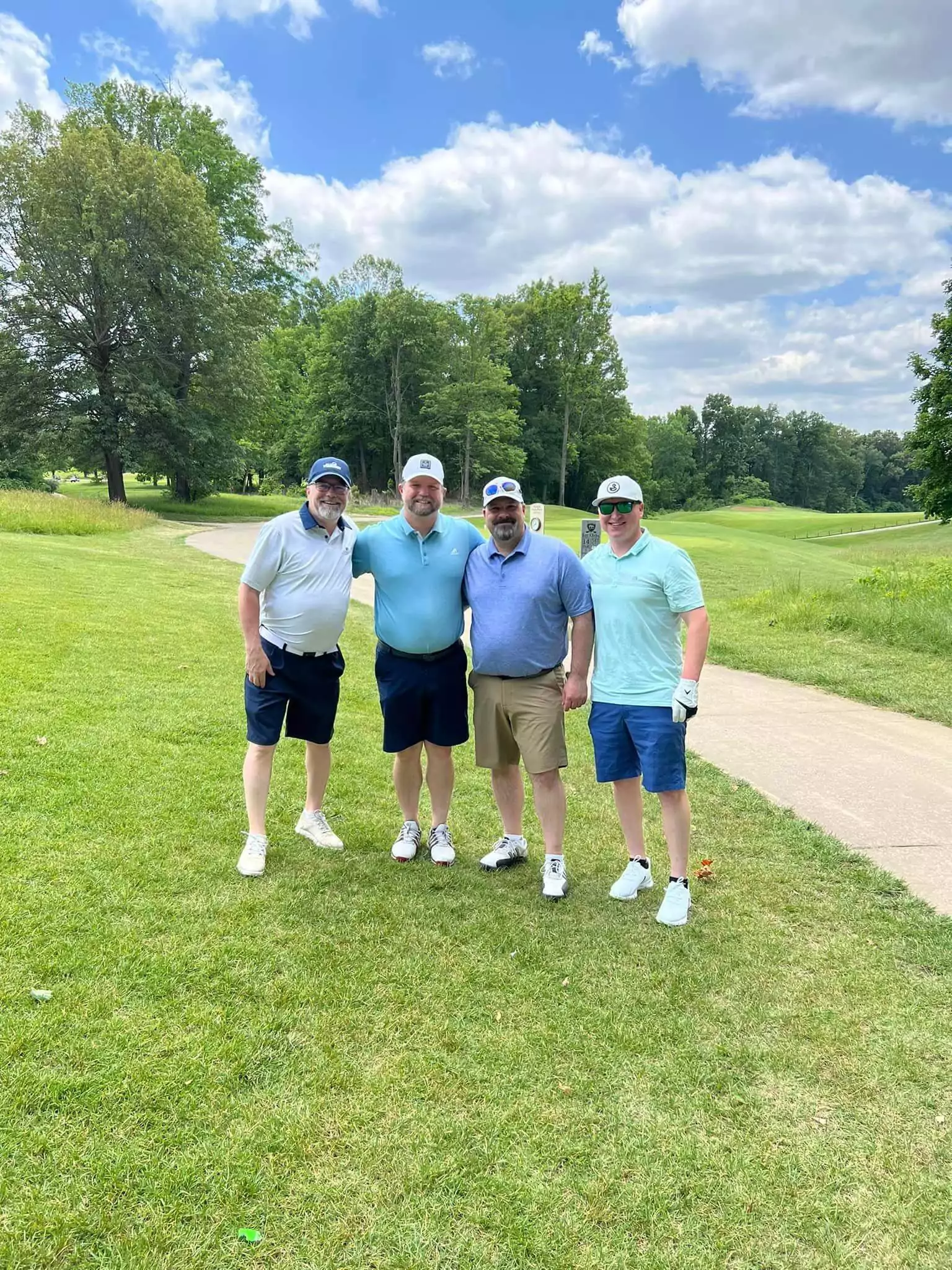 Four men in golf attire stand smiling on a grassy golf course during the SLPCS Spring Golf Tournament 2023. They each wear caps and sunglasses. The sky is partly cloudy, and trees are visible in the background.