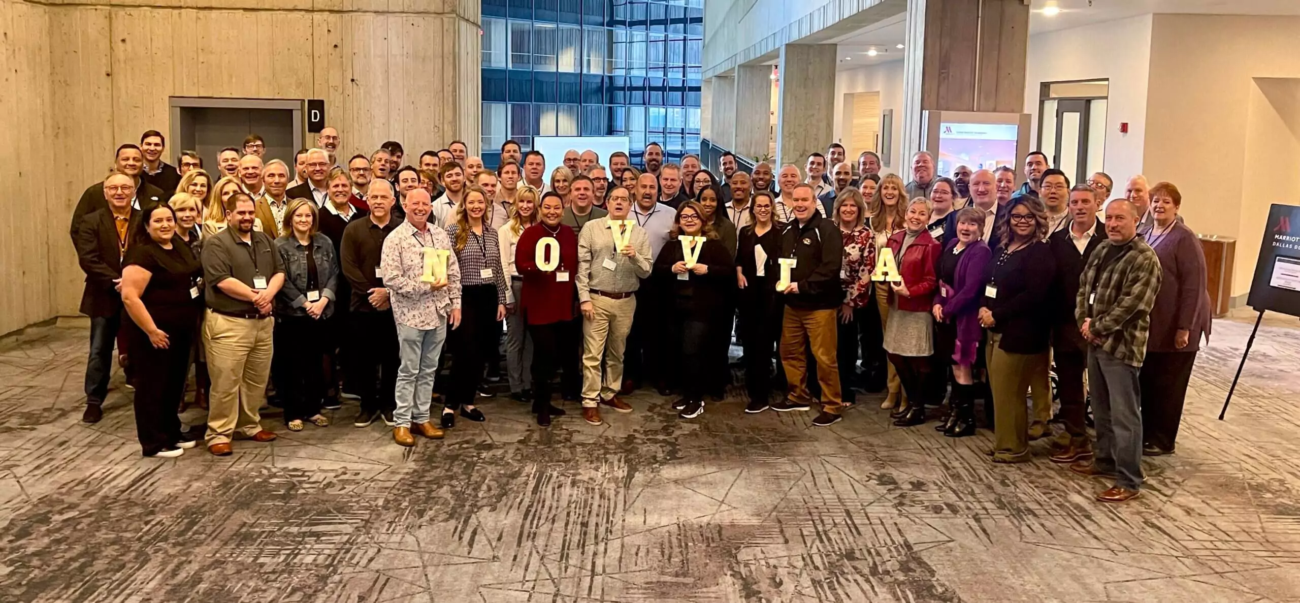 A large group of people smiling and standing closely together in a spacious building with modern architecture. Some individuals are holding large letters that spell "IOVVIA.