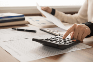 Financial A person uses a calculator on a wooden desk cluttered with documents and a clipboard, evaluating upcoming services. Their other hand holds a sheet of paper. Nearby are a pen and a closed laptop. Sunlight streams from a window in the background.