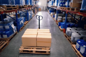 Distributors A pallet jack holds cardboard boxes on a wooden pallet in the center aisle of a warehouse. Shelves filled with blue and white containers line both sides. The warehouse has a polished concrete floor and high ceiling.