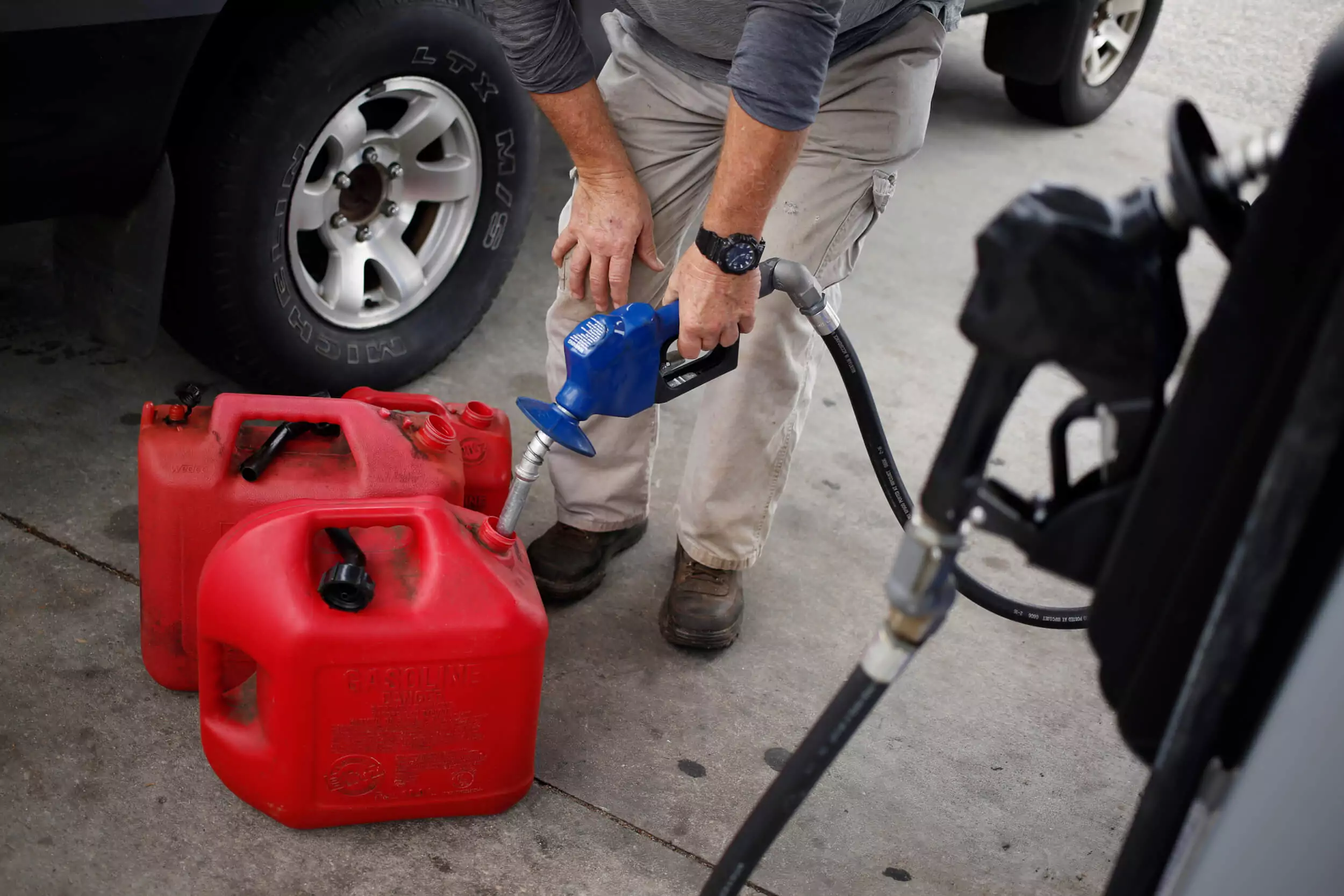 A person in a gray shirt and beige pants refills red portable fuel containers at a gas station using a fuel pump. A vehicle is partially visible in the background.