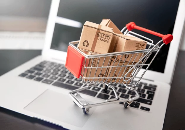 A miniature shopping cart filled with small cardboard boxes sits on a laptop keyboard. The boxes have recycling symbols and the word "fragile" printed on them, representing online shopping or e-commerce.