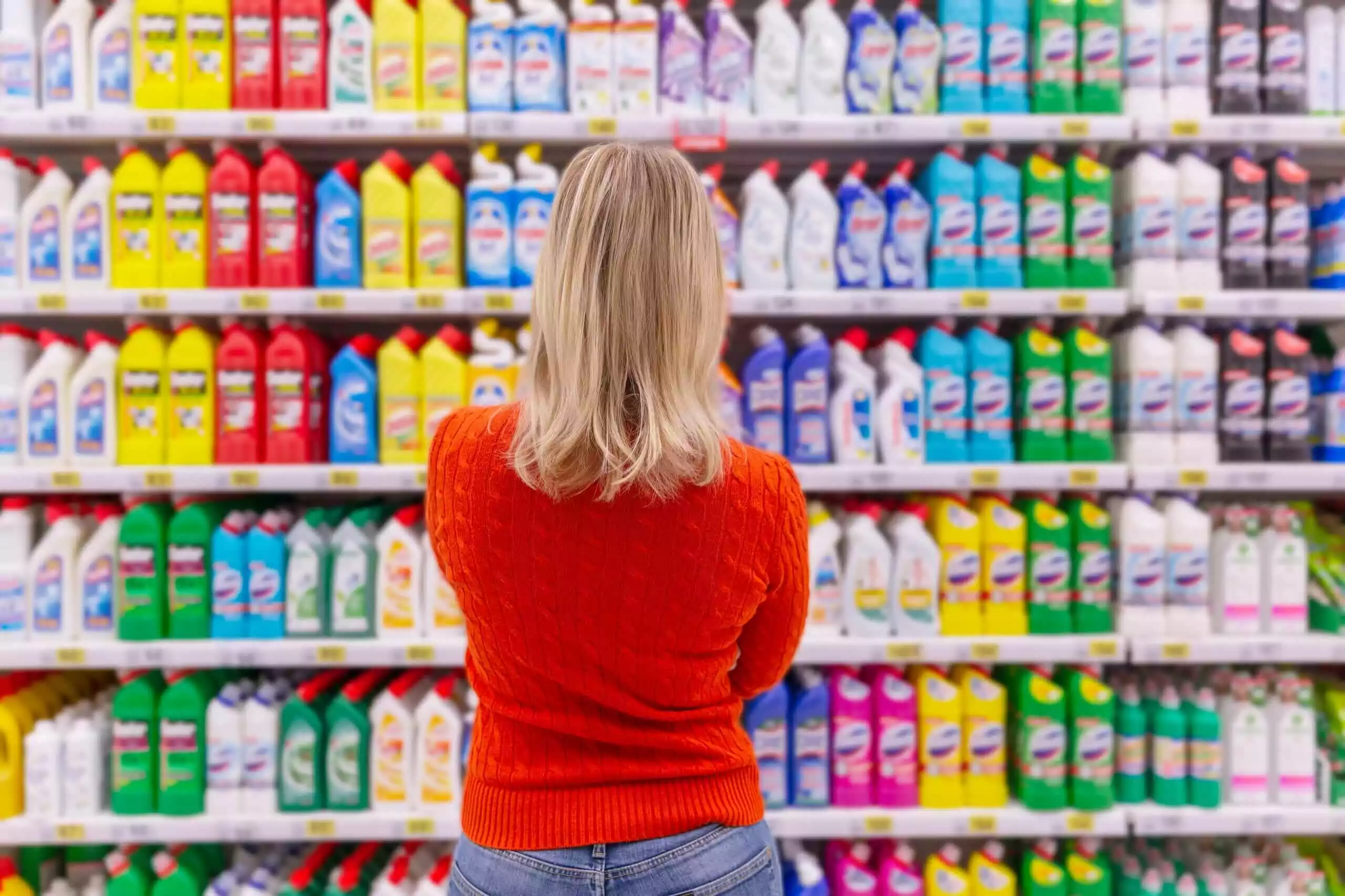A person with blonde hair wearing an orange sweater stands in front of shelves filled with colorful cleaning products in a store. They face the neatly organized shelves, where labels for bottles showcase various brands in different colors and sizes.