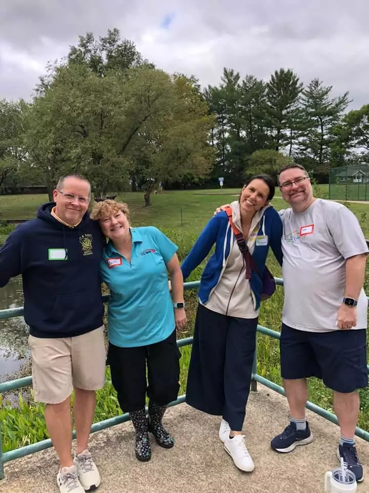 At the Company Picnic 2024, four colleagues stand on a paved path near a railing, smiling warmly. Casually dressed, two lean on each other with camaraderie. Behind them, trees and grass stretch under a cloudy sky, capturing the event's relaxed vibe.