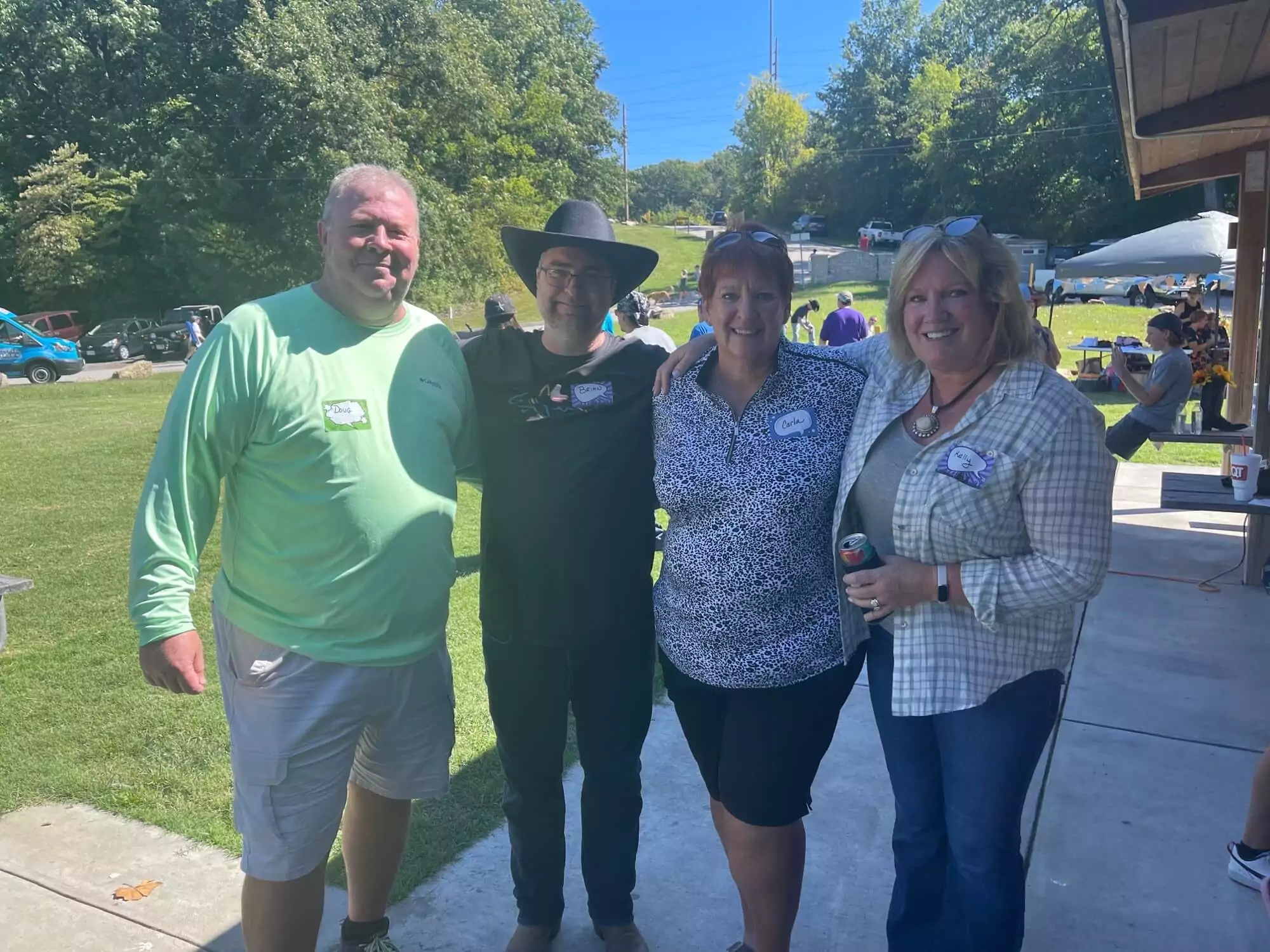 Four people stand together outdoors on a sunny day at the C.L. Smith Company Picnic. They are smiling, with two men on the left and two women on the right, standing on a concrete path surrounded by grass and trees. A picnic table and pavilion are nearby.