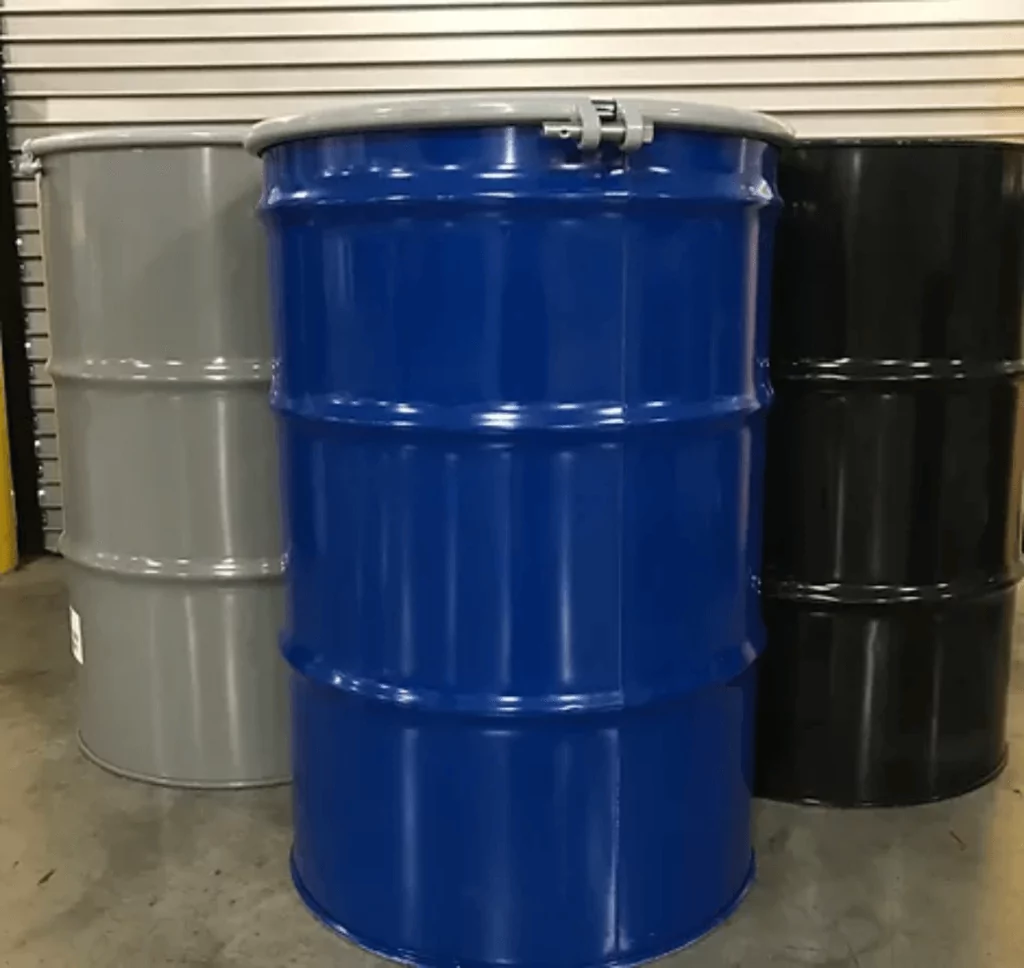 Three metal barrels stand in a row on a concrete floor. The barrels are colored gray, blue, and black from left to right. A metal pipe rests on top of the blue barrel. The background features a closed metal shutter.