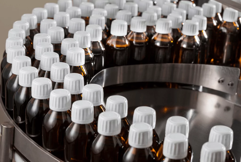 Rows of brown glass bottles with white caps are arranged on a conveyor belt, likely in a manufacturing or packaging facility.