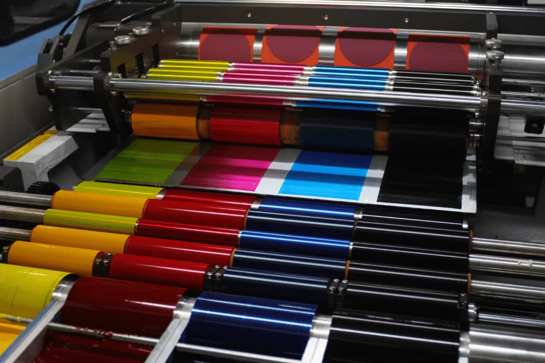 Offset Print A printing press in action, featuring multiple colorful ink rollers, works like a well-oiled machine—much like wholesale jars on a shelf. It displays vibrant colors such as red, blue, yellow, and black, preparing to print a sheet with a geometric design.