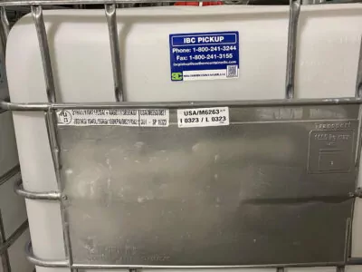 Stacked IBC containers, one prominently labeled with contact info. A barcode and detailed text are visible. Metal cage surrounds the white container, which has a plastic valve at the bottom.