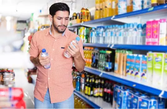 A man wearing a red checkered shirt holds two water bottles while standing in a grocery store aisle. Shelves behind him are stocked with various beverages. He appears to be carefully examining the bottles.