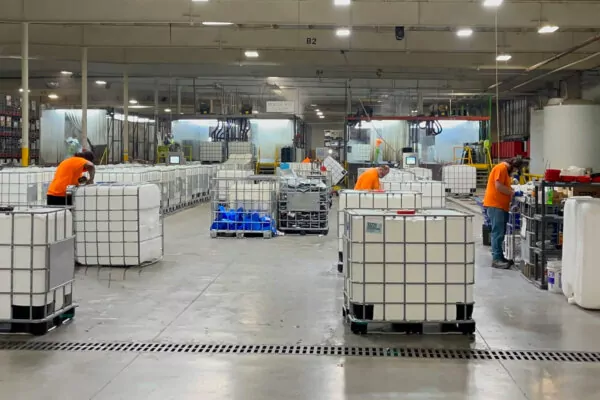 IMG_7031 A spacious industrial facility with three workers wearing orange shirts processing large white containers on pallets. The floor is concrete, and overhead lights illuminate the space. Equipment and shelves line the perimeter.