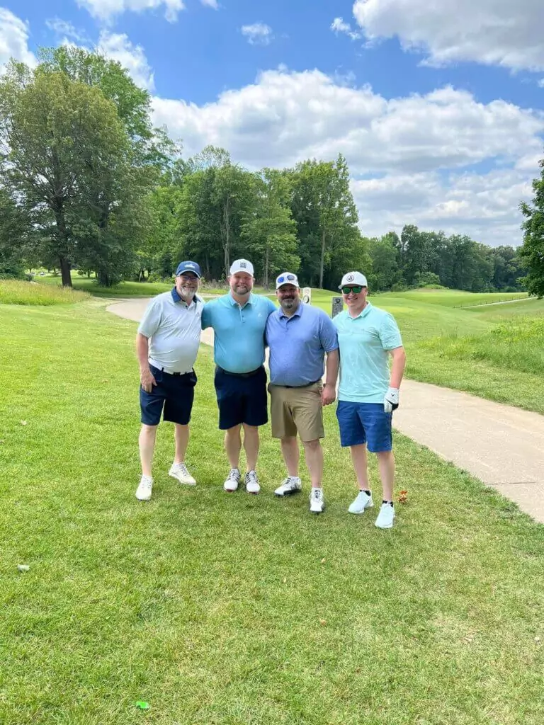 Four men in golf attire stand smiling on a grassy golf course during the SLPCS Spring Golf Tournament 2023. They each wear caps and sunglasses. The sky is partly cloudy, and trees are visible in the background.