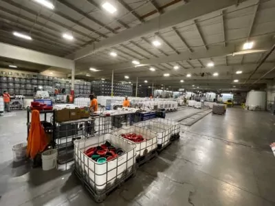 A spacious warehouse with high ceilings, where workers in orange vests are organizing large plastic containers. The floor is glossy, and shelves filled with more containers line the walls. Overhead lighting illuminates the entire area.